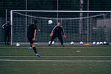 A soccer player in all black practicing shooting on a goalkeeper with orange gloves in goal.