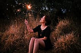A dark-haired woman sitting on a hillside, looking up at the sparkler she’s holding in her hand.