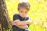 This is an image of a young child with a serious expression, sitting on the grass and leaning against a tree, with arms crossed. The sunlight filtering through the leaves creates a warm, dappled light on the scene.