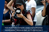 A woman photojournalist wearing a black baseball cap holds up a large camera and lifts her  other arm to hold up the camera’s strap that is around her neck. A message below the image states: For more than 30 years, USAID has supported independent media as part of its democracy promotion efforts. This includes bolstering the professionalism and safety of journalists.