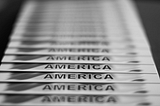 Black and white photo of a row of letter-sized papers with “America” printed at the bottom of each and displayed from foreground to background.