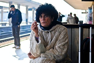 old woman at train station in a sweater with curly black hair smoking a cigarette