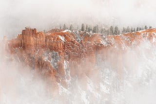 An orange-brown ridge lined with coniferous trees and covered in sections by lightly dusted in snow. Hoodoo rock formations can be seen on the left side of the ridge. The perimeter of the image is largely obscured by fog.