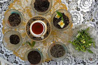 A photograph of six glass containers with loose tea, one with flowers and leaves. There is a cup of tea and saucer in the middle. To the side is a plate of leaves and light green buds. The teacup, containers of tea, leaves and buds are placed on top of a white lace cloth and a gold lace doily.