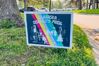 Outside at a park. A sign says, “Nebraska Disability Pride”. White graphics of various disabled people with various disabilities are placed on top of the disability pride flag colors (red, yellow, blue, and greens).