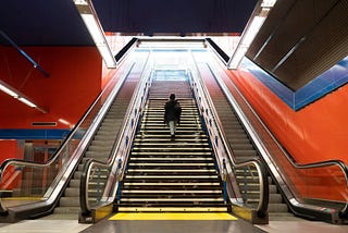 A person ascending an escalator in a modern subway station, flanked by vibrant red and blue walls.