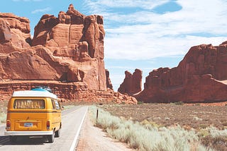 Image of a yellow classic van driving through desert rock formations on a beautiful day.