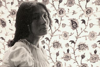 Zitkala-Sa wears a white dress while she sits in front of a flower pattern.