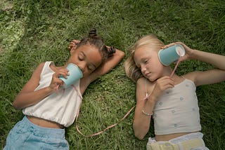 Two young girls laying on grass play telephone with cups and string.