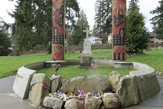 A modest marble cross engraved SEALTH marks the final resting place of Chief Seattle. The cross is butressed by two large cedar columns carved in tribal motifs and painted red and black. The grave site is enclosed by a low cement wall engraved in English and the native dialect with excerpts from a speach attributed to Chief Serattle.