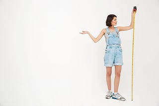 A woman with pale skin holds a tape measure to measure her own height, with a shrugging gesture.