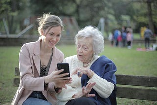 A woman in her 30s helps an elderly woman with something on her phone.