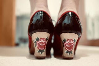 The back of the flamenco shoes of a woman, whose white ankles you can see. The heels are black leather and the heels wooden with red roses and spiky leaves peeping out the sides.