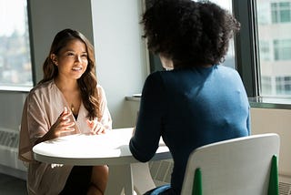 Two women are sat to a table facing each other. One is an young Asian woman talking to another woman that can only seen from the back. Her black curly hair hints that she’s African-American. The picture suggests a coaching relationship.