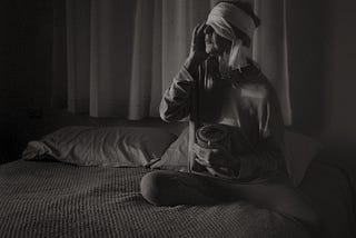 A black and white photo of a woman sitting on a bed with a bandage on her head