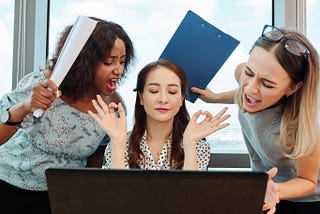 A photo of three women in an office depicting negative work space energy. The boss throws her hands up while two employees become agitated.