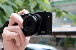 A hand holding a black GR IIIx digital camera, pointing towards the viewer with a blurred background of green foliage and urban streetscape
