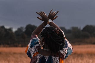 Black womxn with braids in African print dress, back to camera, hands up towards a grey sky.