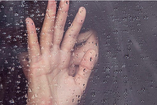 A rain-covered window in front of a hand blocking a woman’s face