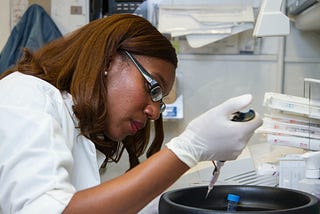 Brown skinned Black womxn in lab coat leans over centrifuge