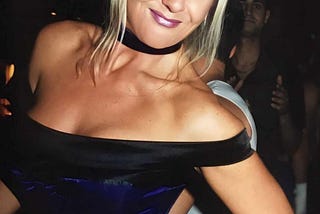 A blonde bombshell woman smiling for the camera. She’s wearing a purple corset and is in a nightclub.