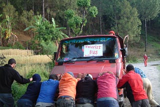 Eight men are pushing the front of a red pickup truck with a sign in the windshield that says “Tourist Bus.” There are trees along the side of the road.