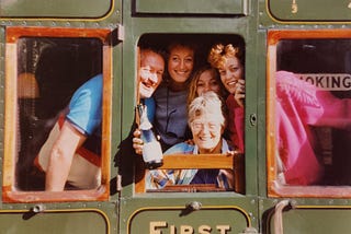 The author’s family of five are inside the first class compartment of an old railway carriage. The author’s mother is holding a bottle of champagne out of the window and everyone is smiling.