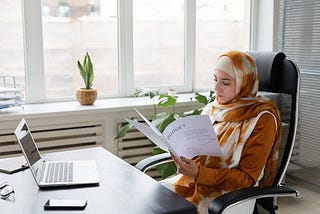 A woman in business attire with a hijab covering her hair in a modern and stylish office sat in front of a computer reading a budget report.