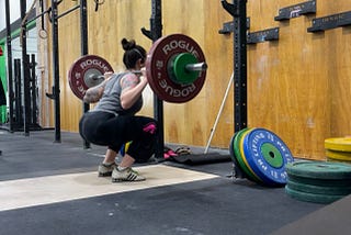 The writer, Kelsey, a white woman, is shown in the bottom of a back squat at a squate rack in a CrossFit gym. She’s wearing weightlifting shoes, leggings, a tank top, and an Aura 3M respirator.