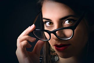 A woman with dark hair and brown eyes lowers her eye glasses