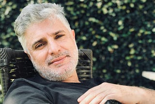 Headshot of a relaxed man with gray hair wearing a black t-shirt sitting outside in a garden with a green bush behind him.