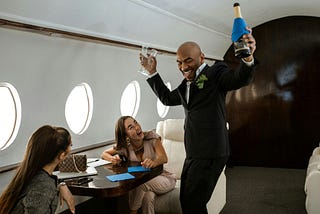 A well-dressed man dancing on a private jet while holding a bottle of champagne and two champagne flutes, with two young women sitting and smiling at him.