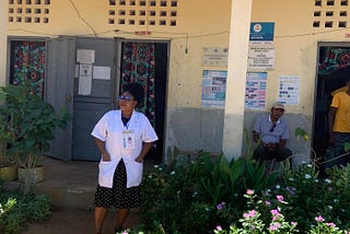 A nurse-midwife wearing a white lab coat stands in front of a community health center with an entrance adorned with lush flowers and greenery.