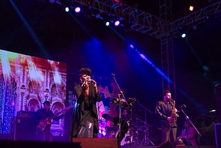A large stage at night lit up with coloured lights, a bass player in the back ground, Black cis female center stage , microphone in hand, young saxophone player to her right.