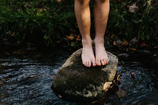 barefoot child stands on a rock in a creek. only the child’s legs, from the knees down, are visible in the photograph