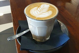 a cup of coffee with latte art in a clear glass along with spoon and saucer.