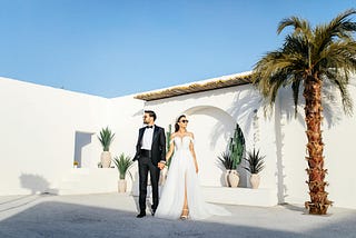 Image shows glamorous bride in wedding gown and groom in tuxedo holding hands and wearing sunglasses outside a bright white casita with blue sky, palm tree, and potted plants