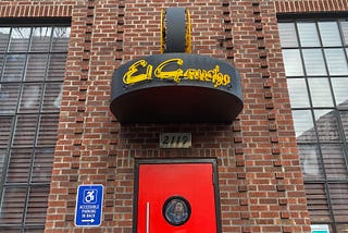 brick building with windows, a red door, and a neon sign that says, “El Gaucho”