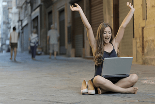 A young woman sitting barefoot in the street with her laptop on her lap, smiling and throwing arms up in a gesture of celebration.