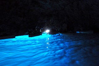 A dark cave with a small point of light in the center behind two rowboats. The water is glowing cerulean blue