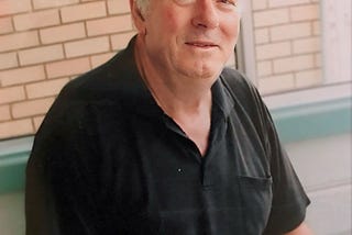 A man in his 70s with grey hair, in a black polo shirt, looking at the camera and smiling slightly.