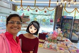 A woman and her daughter, around 15 years old, at a sweet shop. There are hampers of sweets behind them on a large table in the middle of the shop. There are festoons of marigold flowers decorating the shop. A Visicooler with Pepsi in it is at the far end. Another child is in the photo behind the mother and daughter, who is looking at the table with the hampers on it from the far end.