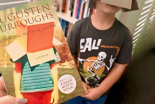 Boy standing in front of a bookshelf with a box on his head, face hidden. A woman holds a book in the forefront, titled, “Running With Scissors” which also has a boy with a box on his head on the front cover.