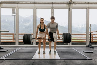 Image shows young woman and man in athletic wear lifting a large barbell together in a gym. The wall behind them is full of windows with view of mountains.