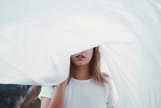 Teenage girl in white partially hiding her face behind a billowing white sheet
