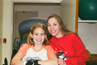 A mom wearing a red sweatshirt with a snowman. The mom has her arm around her ten-year-old daughter who is in a standing frame at the hospital.
