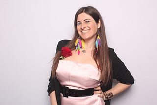 The author, me, dressed in a smart outfit composed of a satin baby pink dress and a black vest complimented with  multicolored feather earrings.