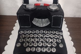 A cake in the design of an old black typewriter with silver keys and piece of white paper made of frosting with the writing Happy Birthday JoEllen!