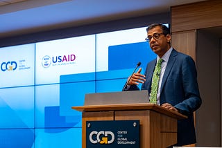 USAID Assistant Administrator for Global Health Dr. Atul Gawande speaks into a microphone while standing behind a podium adorned with the logo CGD. Behind him is an electronic display with the logos of CGD and USAID.