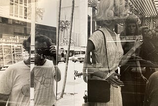 There are two images in this photography: 1) On the right side, there a mannequins inside a store, and 2) On the left side, the split images of the photographer and the city streets and building behind him.